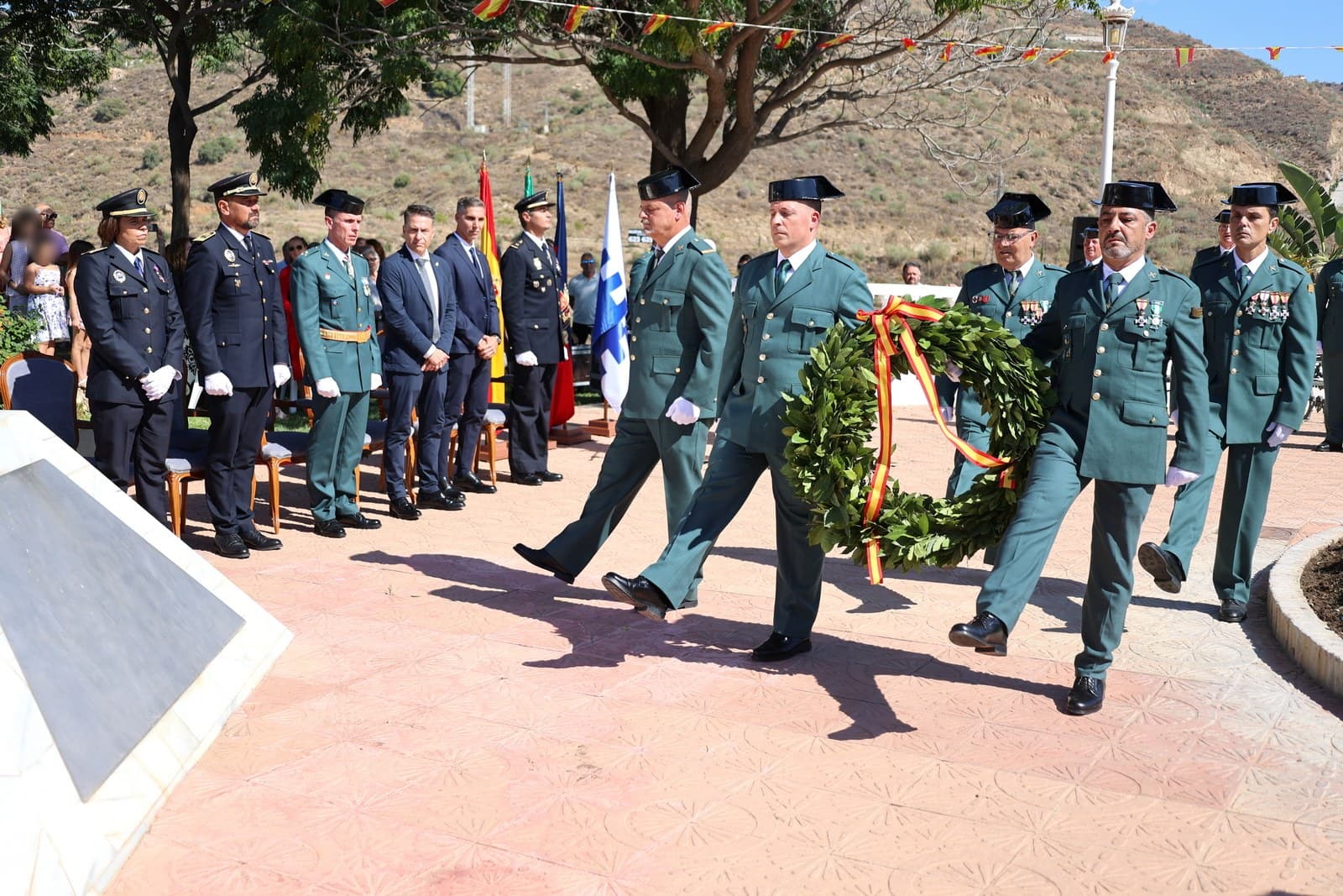Cuatro guardias civiles con tricornio y uniforme de gala llevan una corona de laurel con una cinta roja y amarilla, mientras policías locales, guardias civiles y autoridades los observan en una plaza de Torrox
