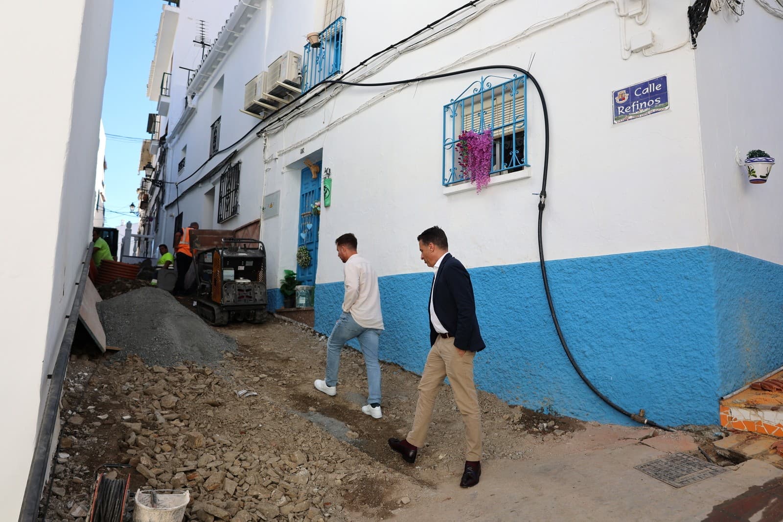 El alcalde de Torrox y un concejal suben por la calle Refinos, con el suelo levantado, una máquina y operarios trabajando en la zanja entre casas blancas con zócalo azul.