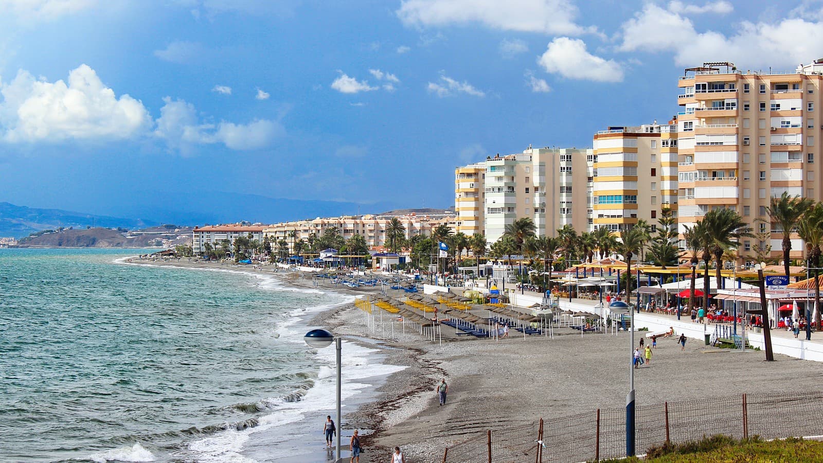 Paseo marítimo y playa de Torrox Costa, con la arena gris, las sombrillas, los bloques de pisos de la primera línea entre palmeras y el mar a la izquierda bajo un cielo con nubes.