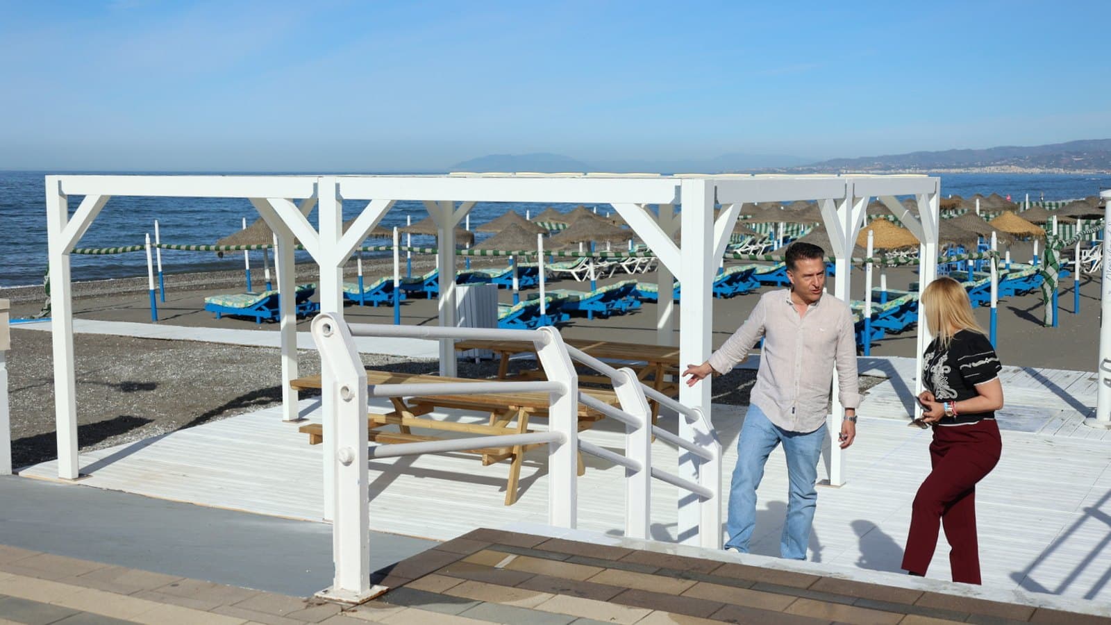 Un hombre y una mujer en una pasarela de madera blanca con pérgola y mesas de picnic en una playa de Torrox, con hamacas azules, sombrillas de paja y el mar al fondo