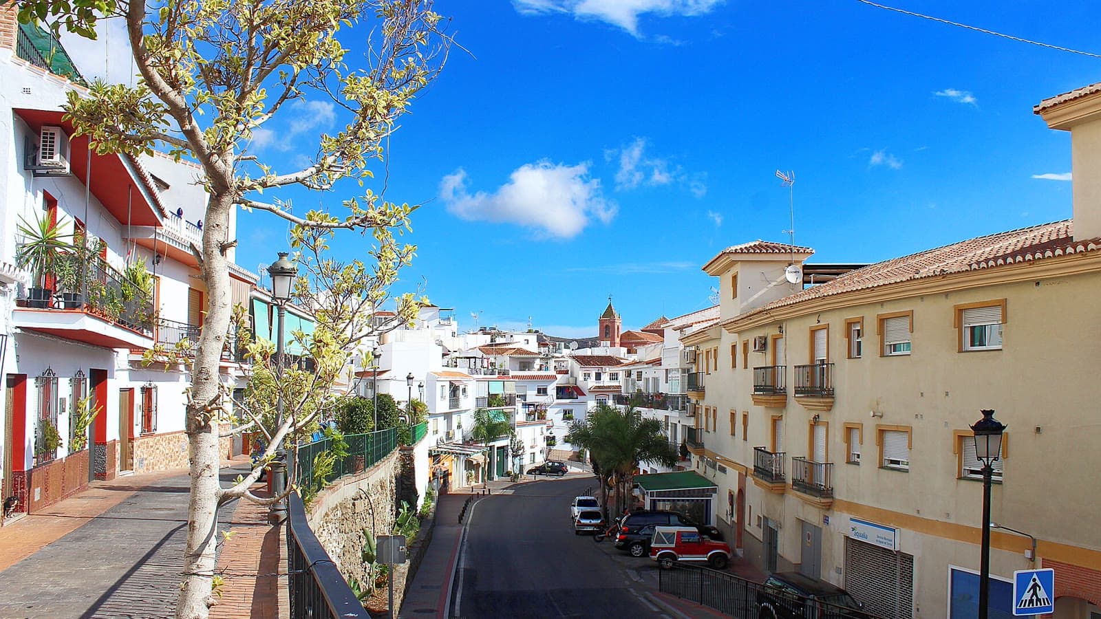 Calle de Torrox Pueblo con casas blancas y balcones, coches aparcados junto a la acera y, al fondo, un campanario sobre los tejados, bajo un cielo azul con nubes.