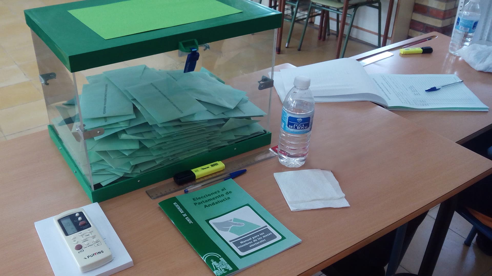 Urna transparente llena de sobres sobre la mesa de un colegio electoral, junto al manual de los miembros de mesa