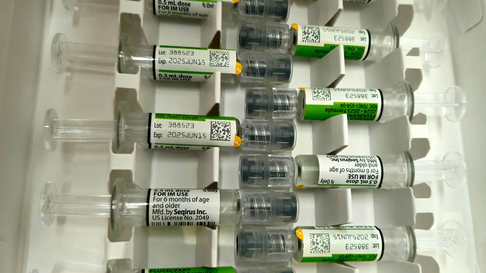 Caja abierta con diez jeringas precargadas de vacuna antigripal, con etiquetas verdes y blancas, colocadas en dos filas sobre un soporte de cartón blanco.