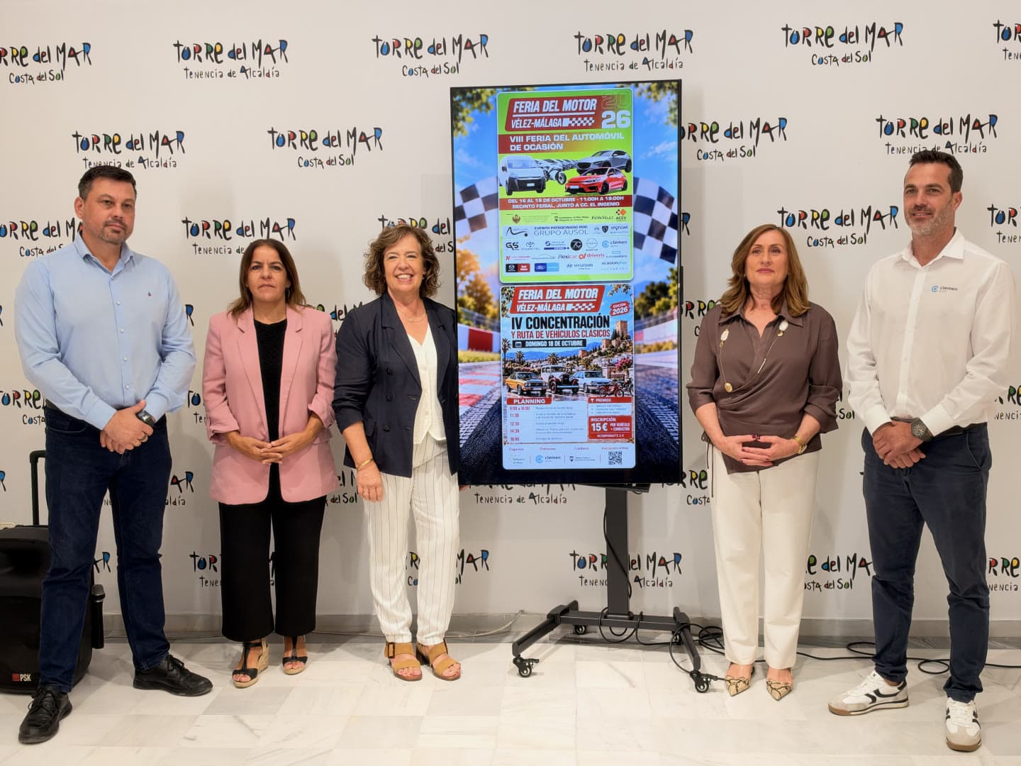 Cinco personas posan junto a una pantalla con los carteles de la Feria del Motor de Vélez-Málaga 2026 ante un photocall de la Tenencia de Alcaldía de Torre del Mar