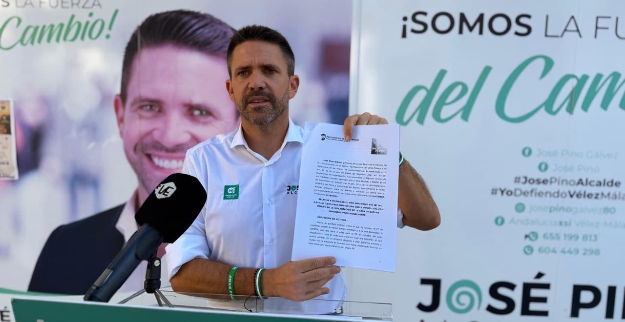 José Pino, con camisa blanca y la pulsera verde de Andalucía Por Sí, enseña ante un micrófono el escrito de su propuesta para modificar el tipo del IBI; detrás, carteles de su candidatura.