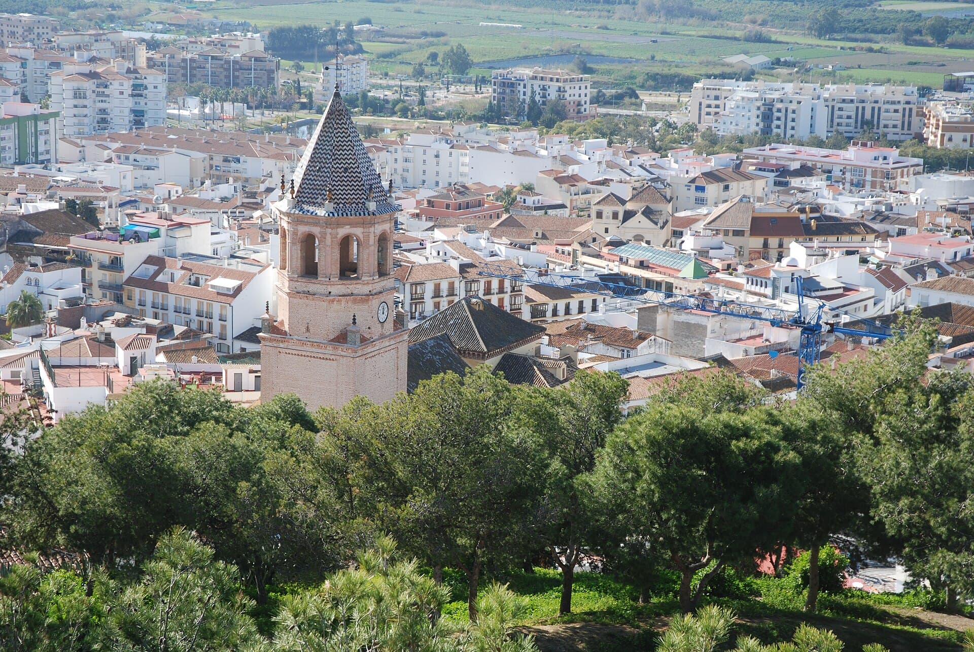 Casas y tejados de Vélez-Málaga vistos desde la fortaleza, con el campanario de la iglesia de San Juan Bautista en el centro y edificios de pisos al fondo.