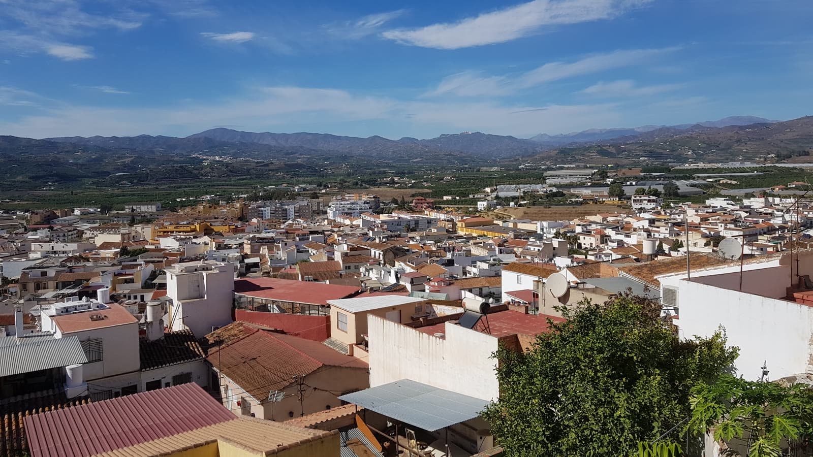 Vista desde lo alto de los tejados y casas blancas de Vélez-Málaga, con la vega, los invernaderos y las sierras de la Axarquía al fondo bajo un cielo azul.