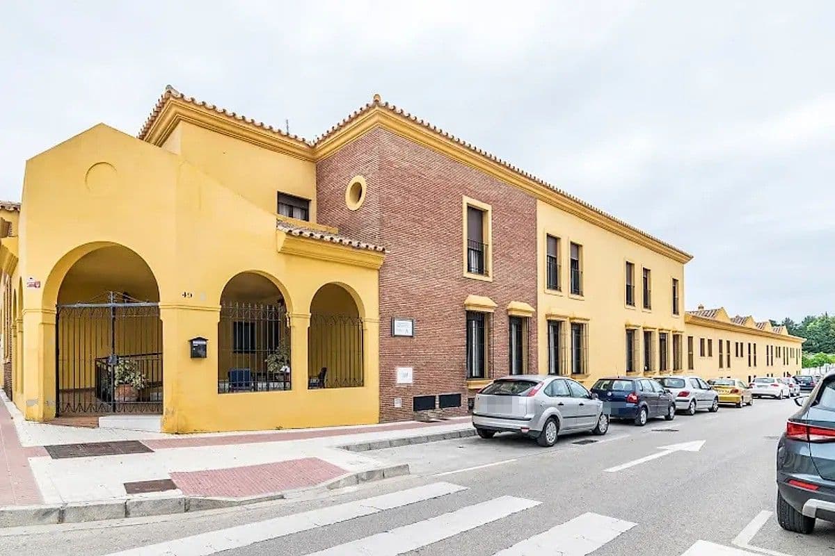 Exterior de la residencia de mayores Seniors desde la calle: un edificio alargado de dos plantas, con fachada amarilla y de ladrillo, arcos con rejas en la entrada y coches aparcados delante.