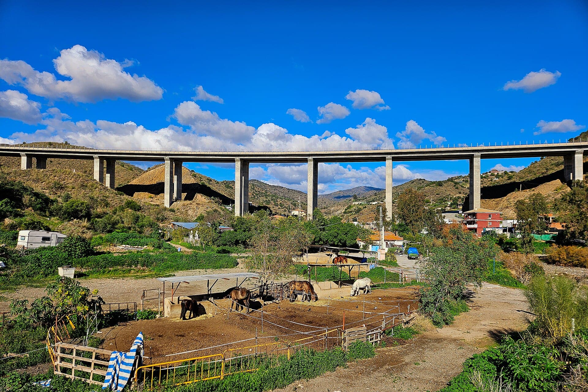 Viaducto de la autovía A-7 sobre el arroyo Totalán, entre lomas verdes y un cielo azul con nubes, con casas y huertos al pie.