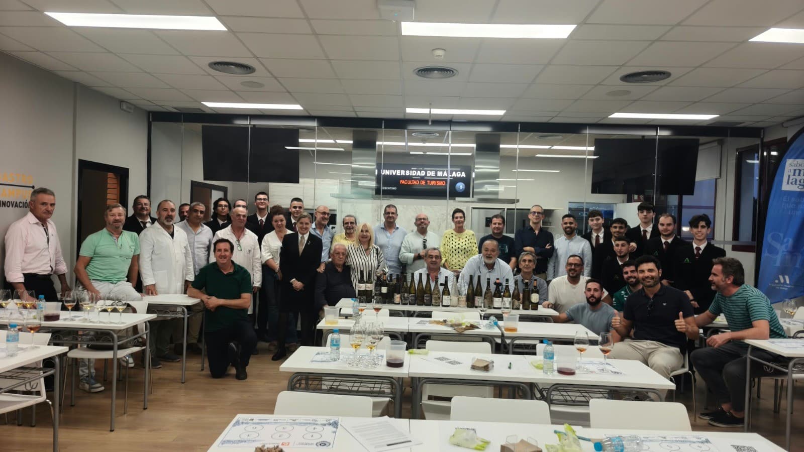 Foto de grupo de unas cuarenta personas en el laboratorio de la Facultad de Turismo de la Universidad de Málaga, con botellas de vino alineadas en una mesa y copas de cata en primer plano.