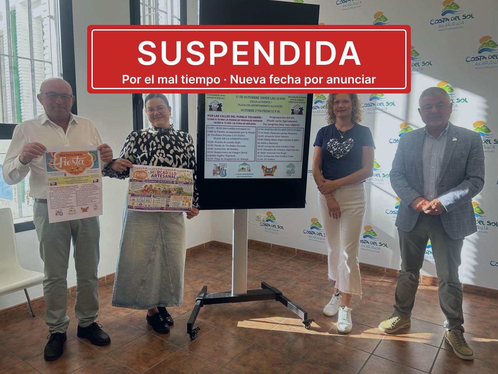 Presentación de la Fiesta Multicultural de Alcaucín en la Mancomunidad: cuatro personas junto a una pantalla con el programa y dos carteles, con un rótulo rojo superpuesto que dice «Suspendida. Por el mal tiempo. Nueva fecha por anunciar».