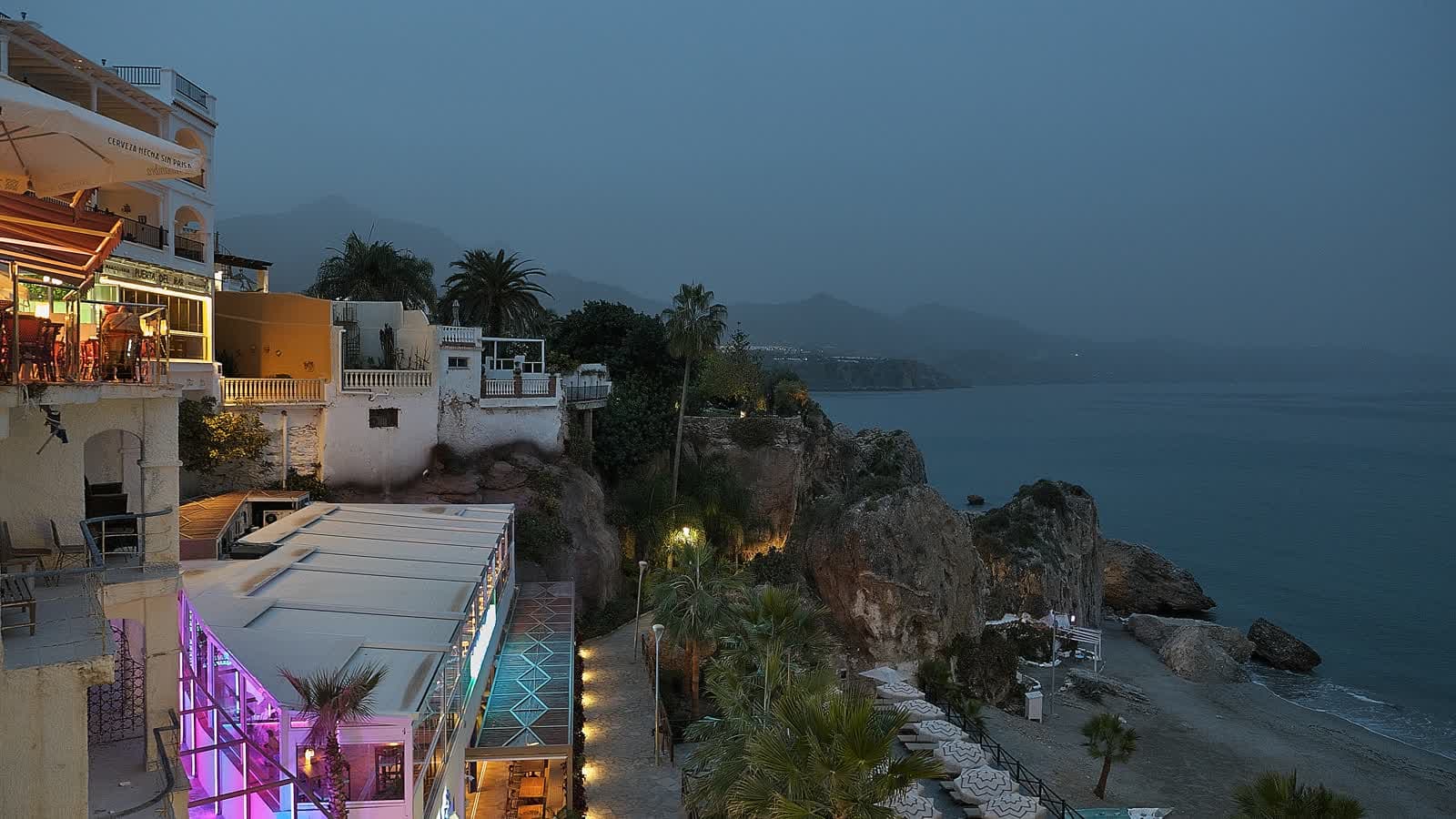 Anochecer en Nerja: edificios iluminados sobre el acantilado, palmeras y una playa junto al mar en calma, con las montañas al fondo.