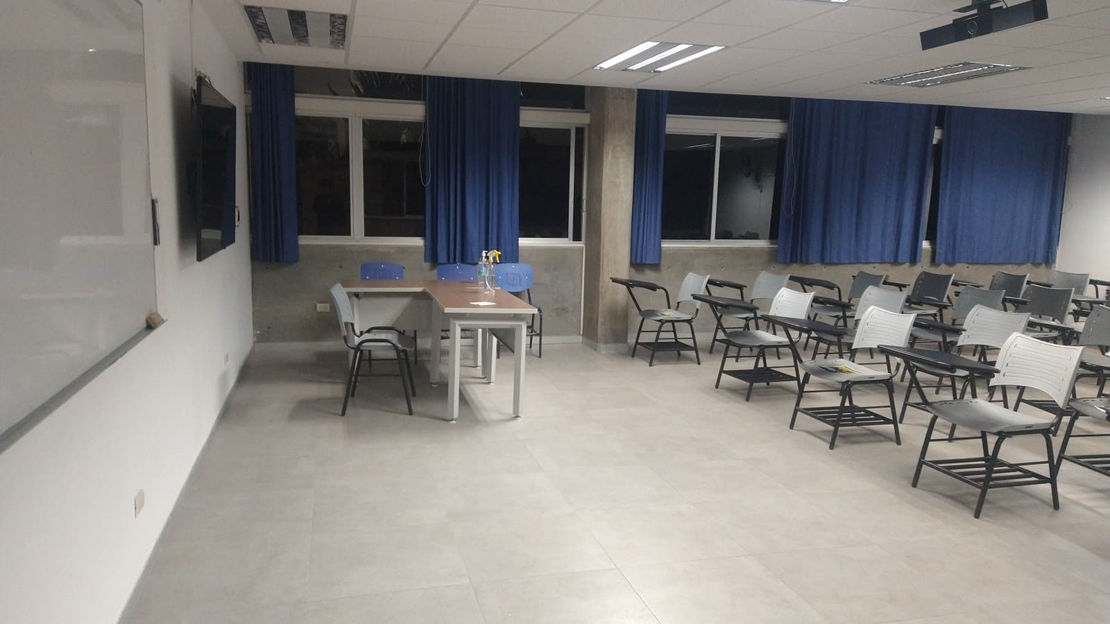 Aula vacía con sillas de pala grises en filas, una mesa de profesor, pizarra blanca y cortinas azules en las ventanas.