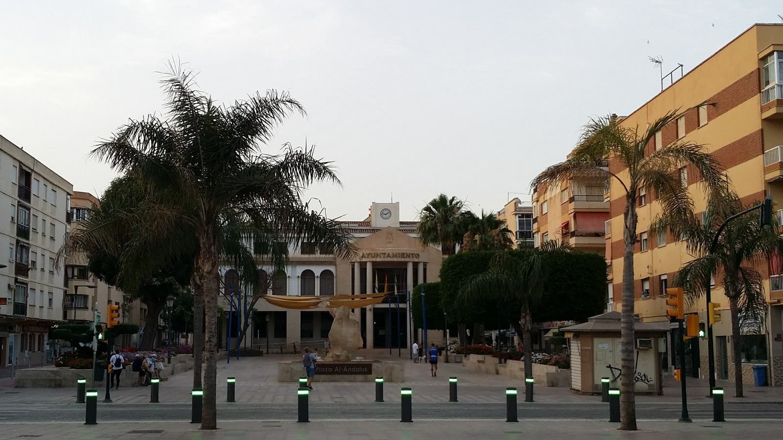 Plaza Al-Ándalus de Rincón de la Victoria con palmeras, una escultura en el centro y, al fondo, el edificio del Ayuntamiento con su reloj.