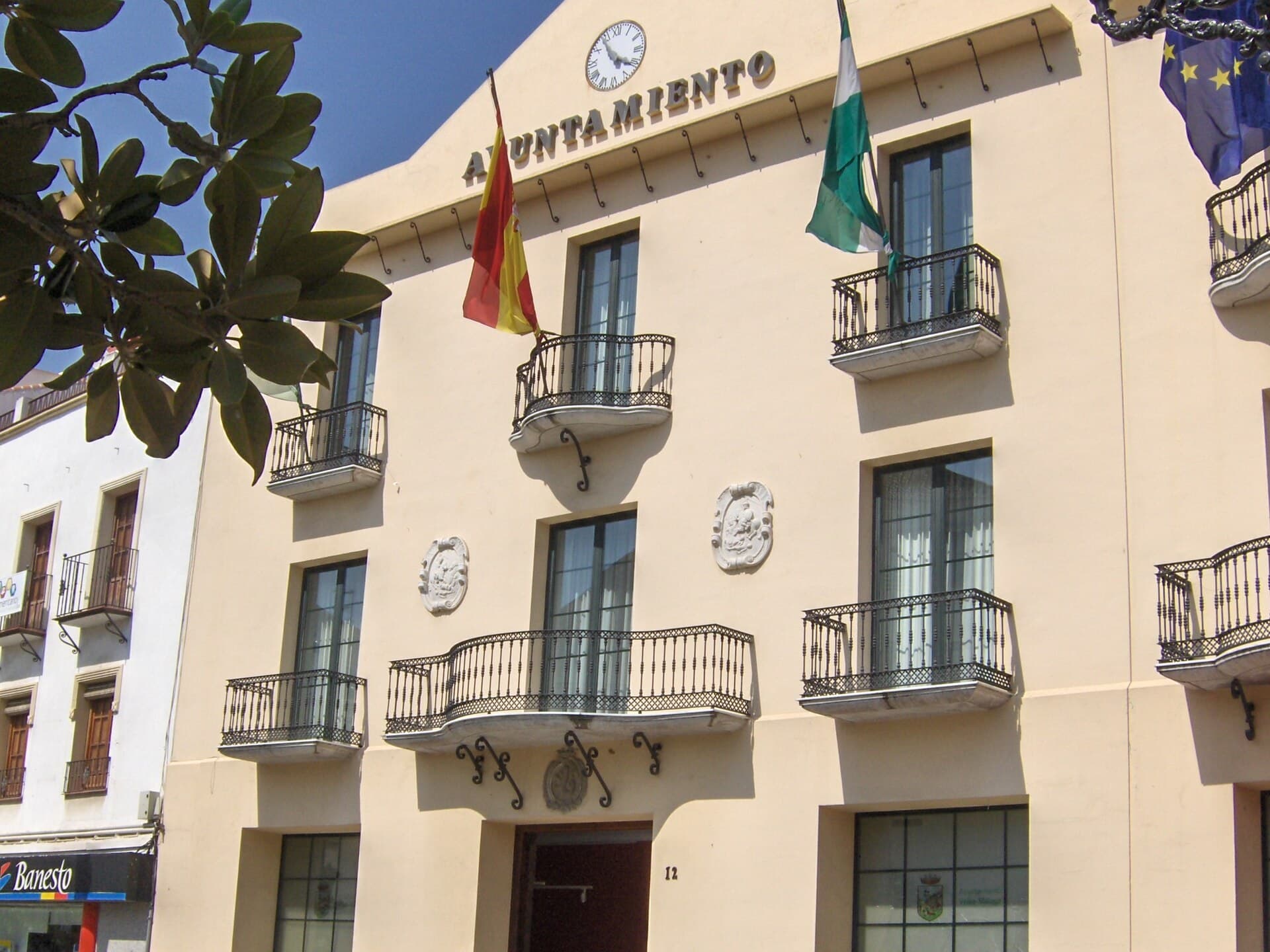 Fachada del Ayuntamiento de Vélez-Málaga, de color crema, con reloj, balcones de forja y las banderas de España, Andalucía y la Unión Europea.