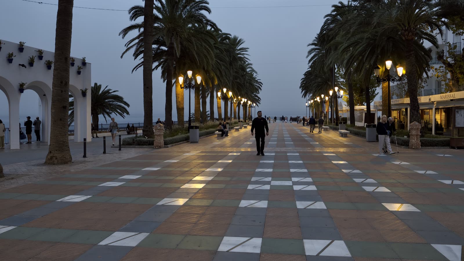 Paseo del Balcón de Europa de Nerja al anochecer, con suelo de baldosas de colores, dos hileras de palmeras, farolas encendidas y algunos paseantes.