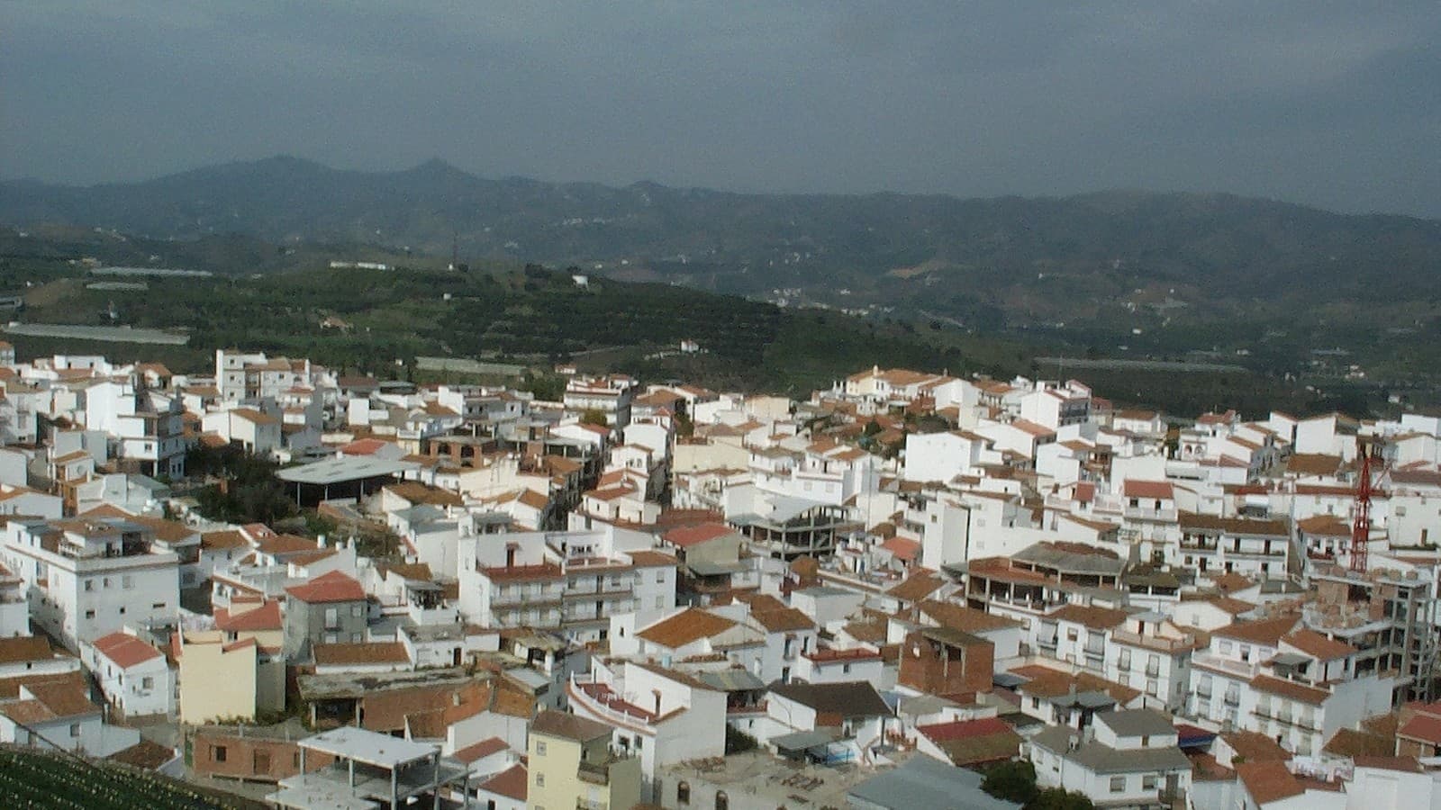 Vista panorámica de Benamocarra desde el Cerro de la Jaula: un caserío de casas blancas con tejados rojos y, al fondo, campos de cultivo y montañas bajo un cielo nublado.