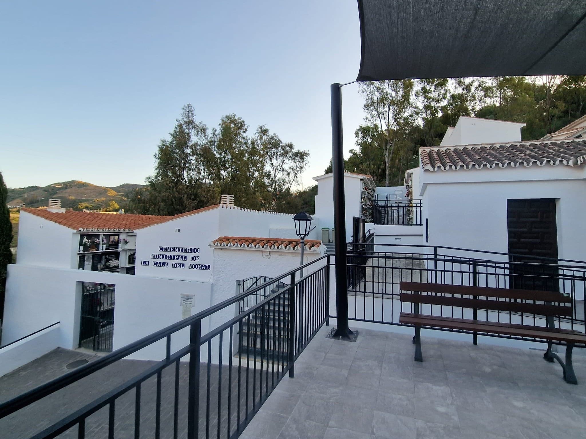 Entrada encalada del cementerio municipal de La Cala del Moral, con su nombre en azulejos, tejados de teja y una terraza con barandilla y un banco a la sombra de un toldo.