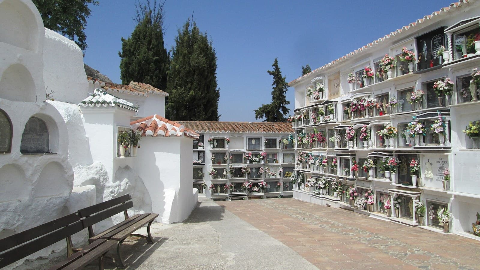 Patio del cementerio de Comares: a la derecha, un bloque de nichos encalados con flores de colores y tejadillo de teja; a la izquierda, antiguas sepulturas blancas de formas redondeadas y dos bancos de madera, con cipreses al fondo bajo un cielo azul.