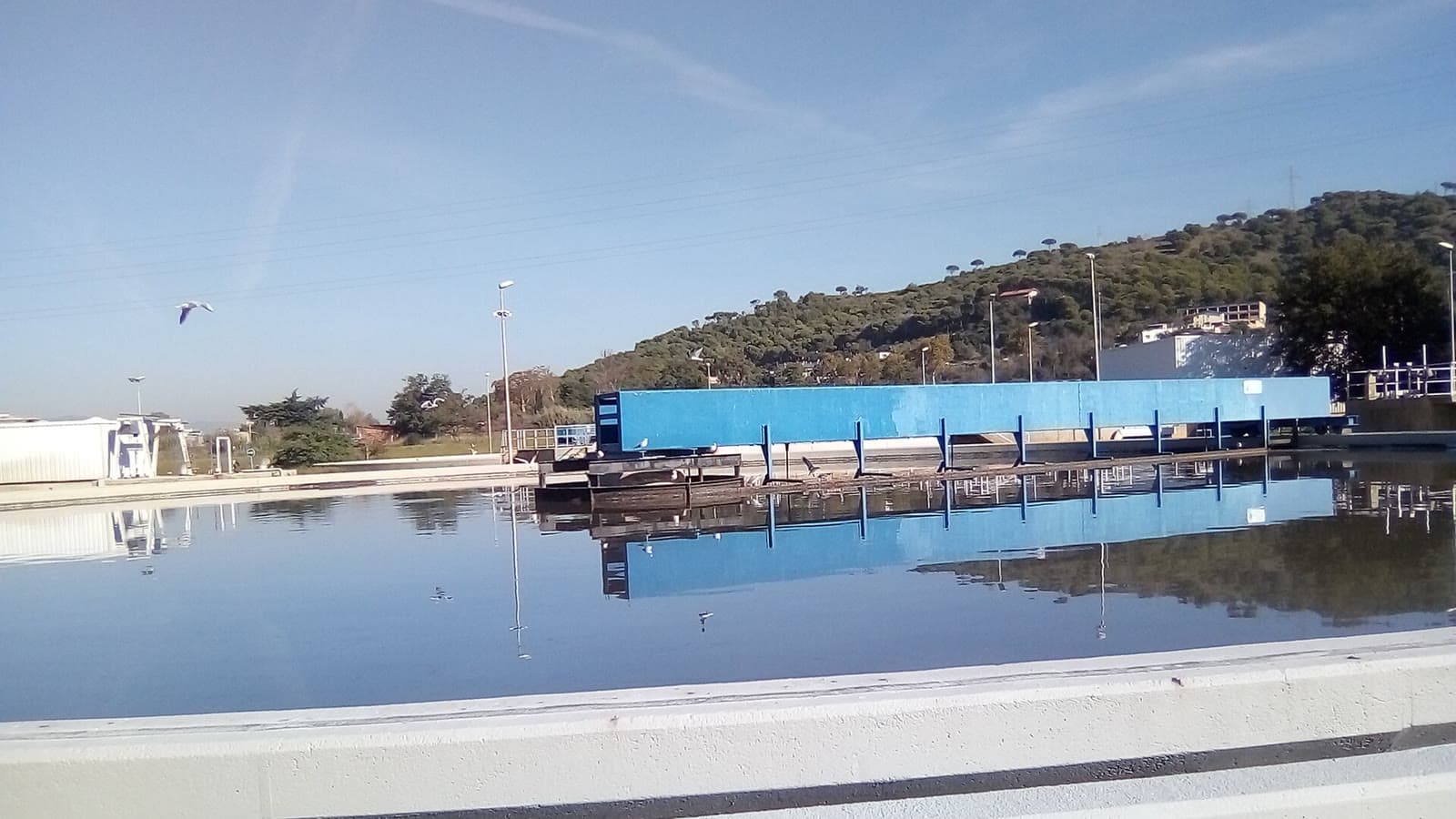 Decantador de una estación depuradora de aguas residuales: una gran balsa de agua quieta con un puente azul que la cruza y una ladera con árboles al fondo.