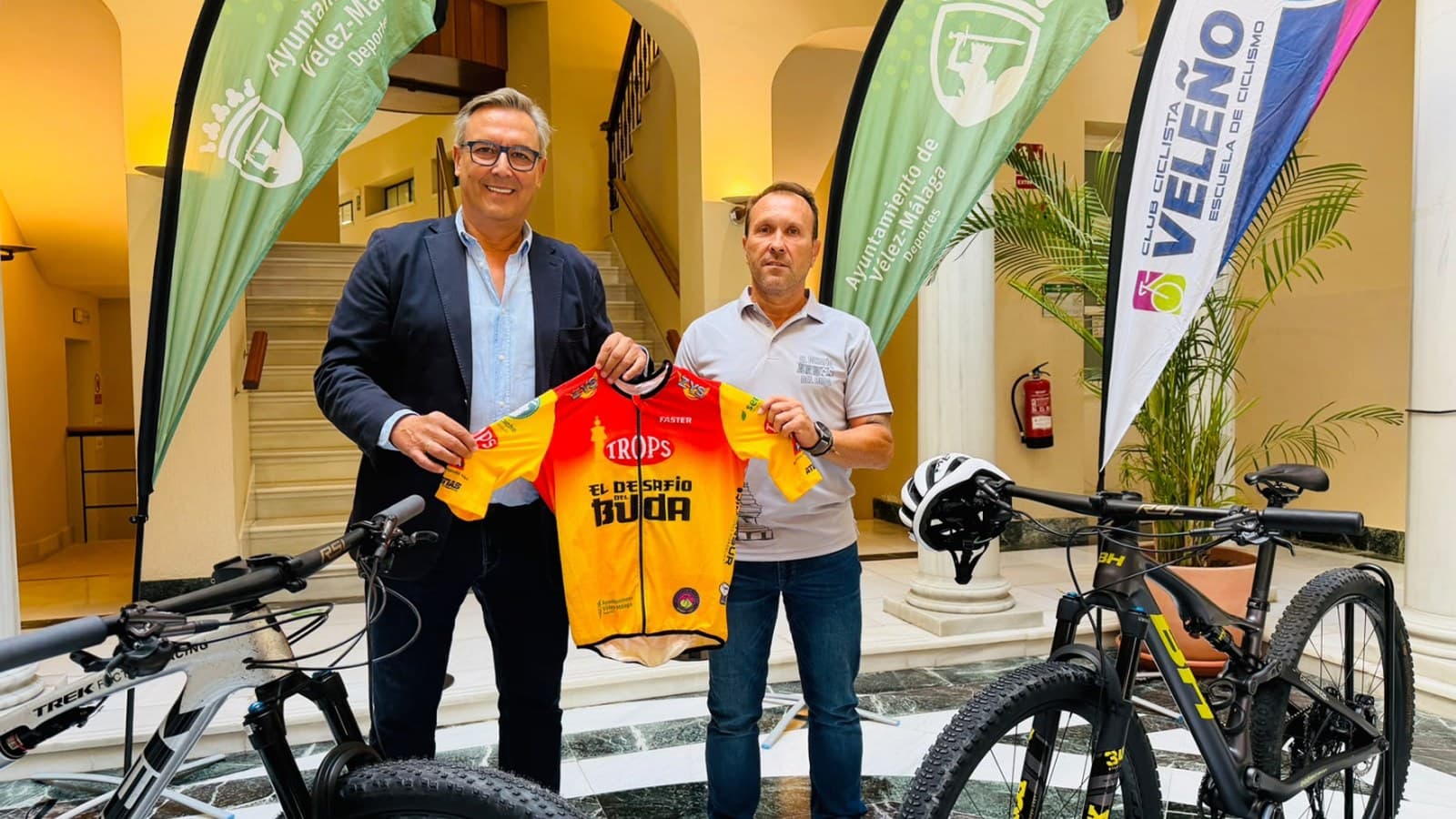 Dos hombres sostienen un maillot amarillo y rojo con el texto «El Desafío del Buda» en un patio con escalera y columnas, entre dos bicicletas de montaña y banderolas del Ayuntamiento de Vélez-Málaga y del Club Ciclista Veleño.
