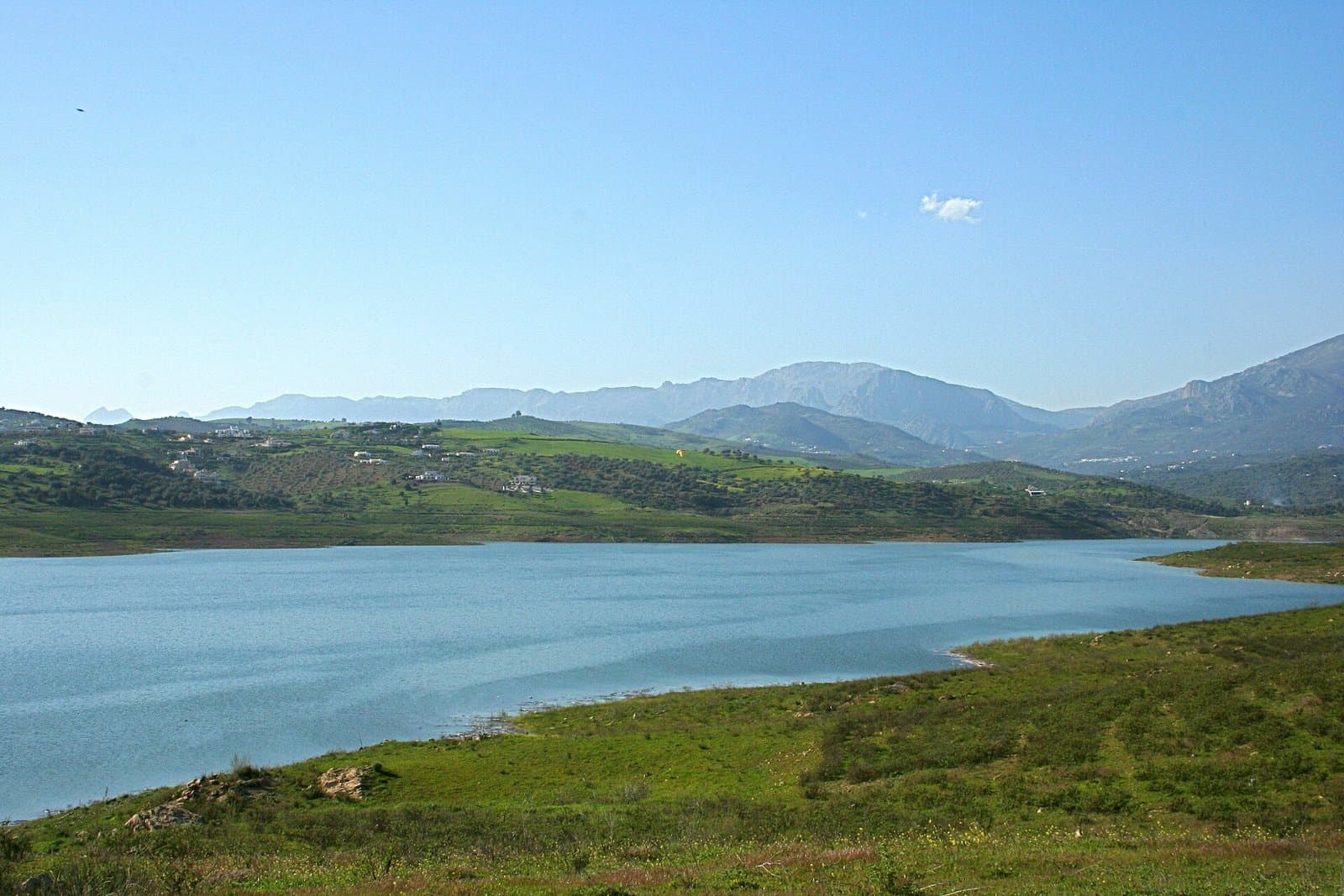 Embalse de La Viñuela con el agua alta, rodeado de laderas verdes y con la sierra al fondo