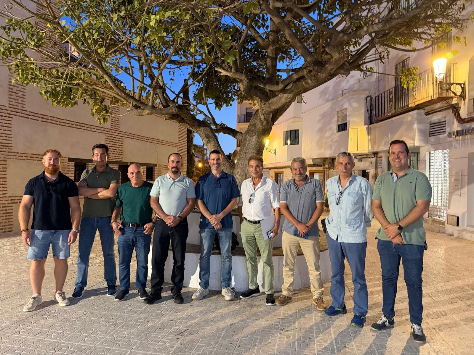Nueve hombres posan juntos en una plaza de Nerja, bajo un gran árbol, al anochecer