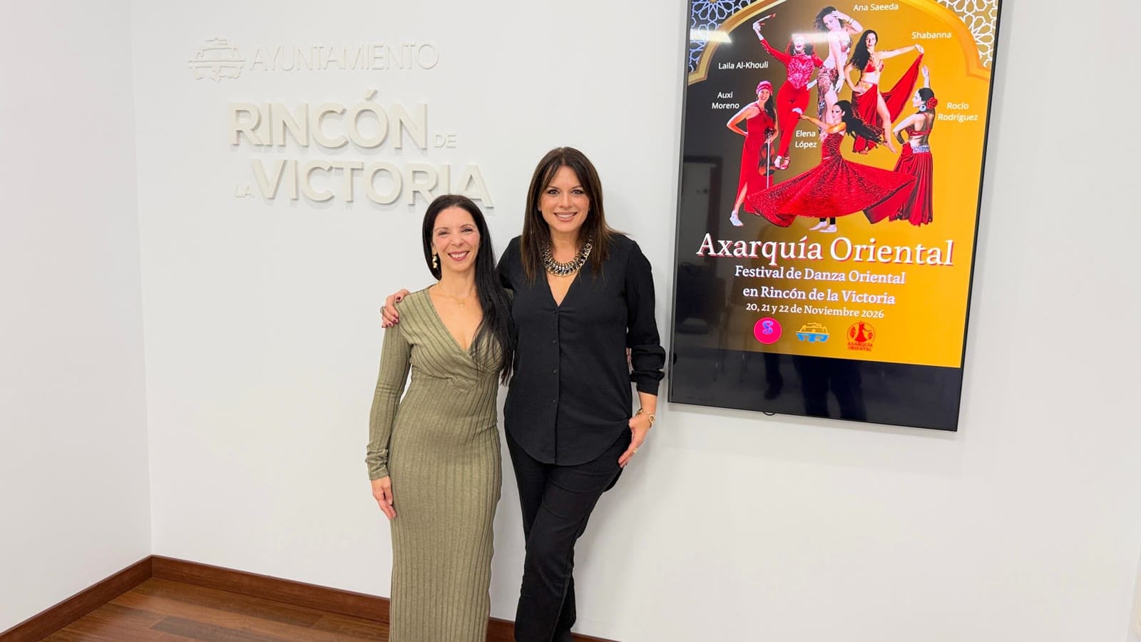 Dos mujeres posan en una sala del Ayuntamiento de Rincón de la Victoria junto a una pantalla con el cartel amarillo y rojo del festival Axarquía Oriental, con varias bailarinas de danza oriental.