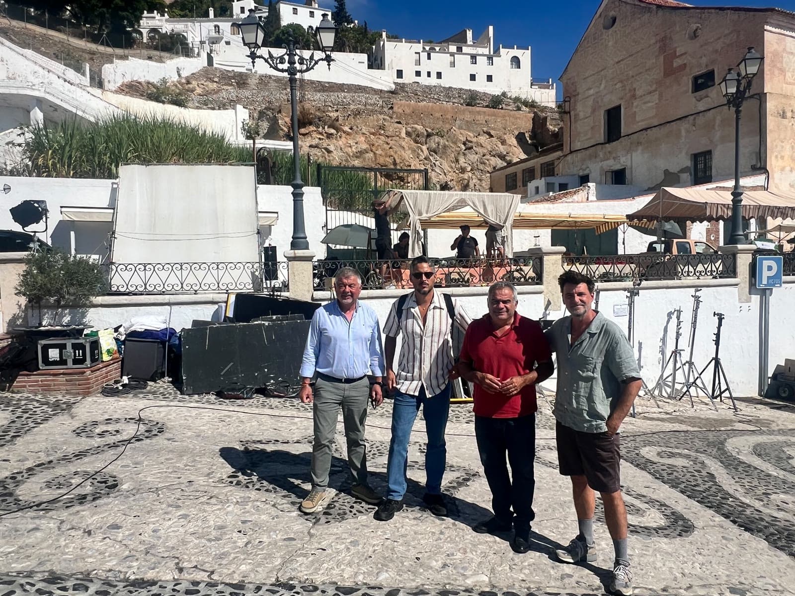 Cuatro hombres posan en una plaza empedrada de Frigiliana junto a focos, trípodes y material de rodaje, con casas blancas y una ladera rocosa al fondo