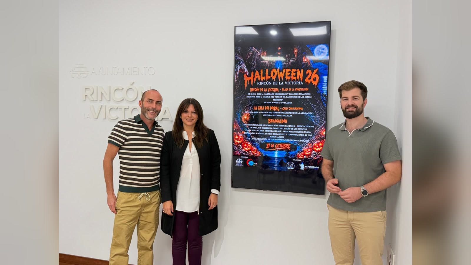 Dos hombres y una mujer posan sonrientes junto a una pantalla vertical que muestra el cartel de Halloween 26 de Rincón de la Victoria, con calabazas, una luna llena y el programa por lugares, en una sala del Ayuntamiento con el rótulo «Ayuntamiento Rincón de la Victoria» en la pared.