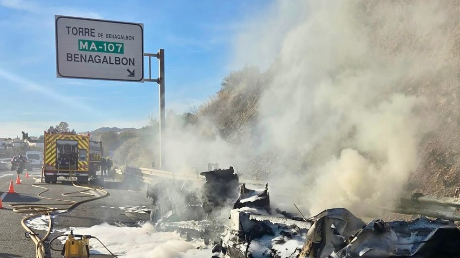 Restos calcinados de una autocaravana en el arcén de la A-7, cubiertos de espuma y humo, con un camión de bomberos detrás y el cartel de salida a Torre de Benagalbón y Benagalbón (MA-107).