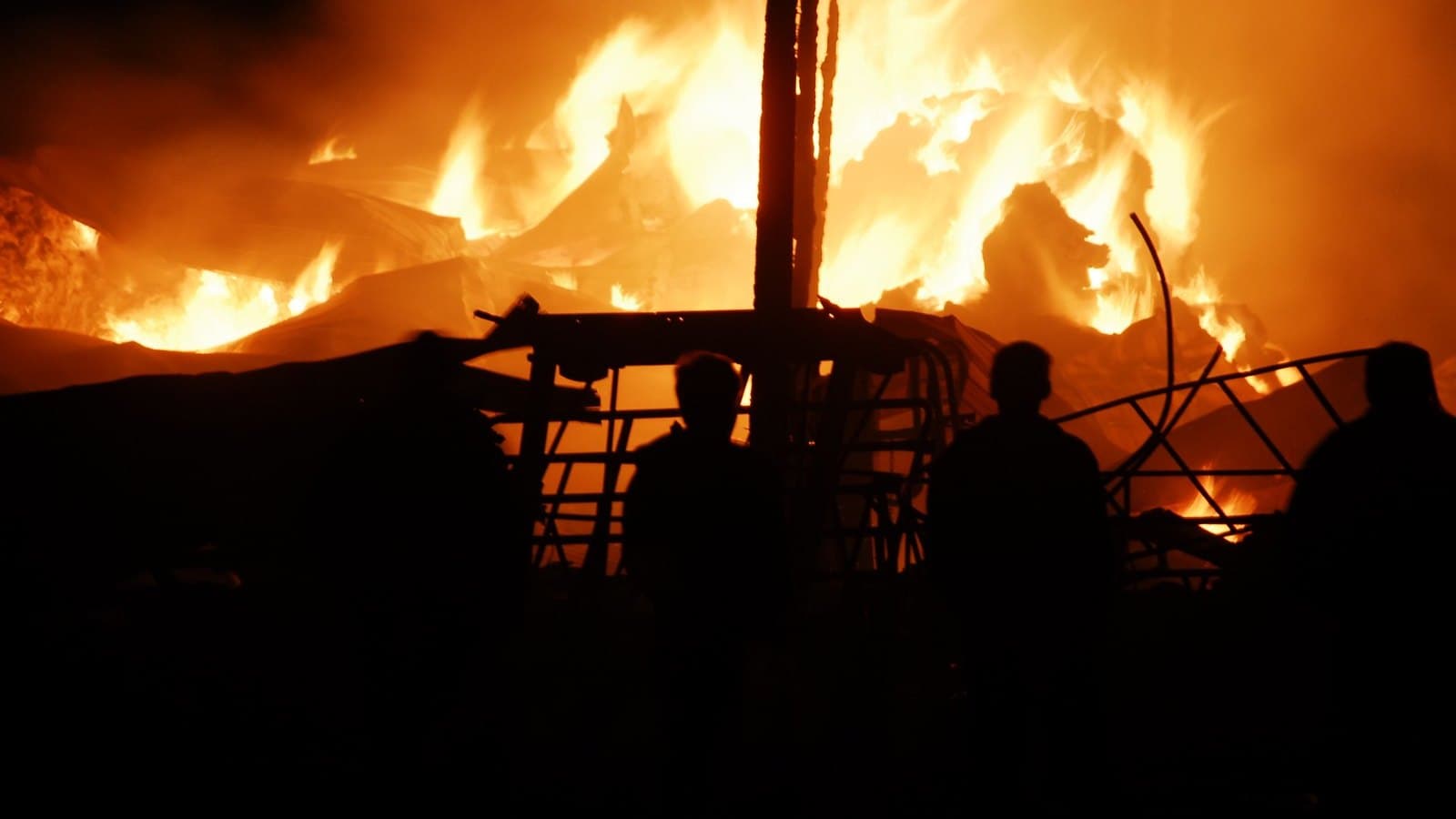 Llamas consumiendo de noche una construcción de madera y chapa, con varias personas a contraluz frente al fuego.