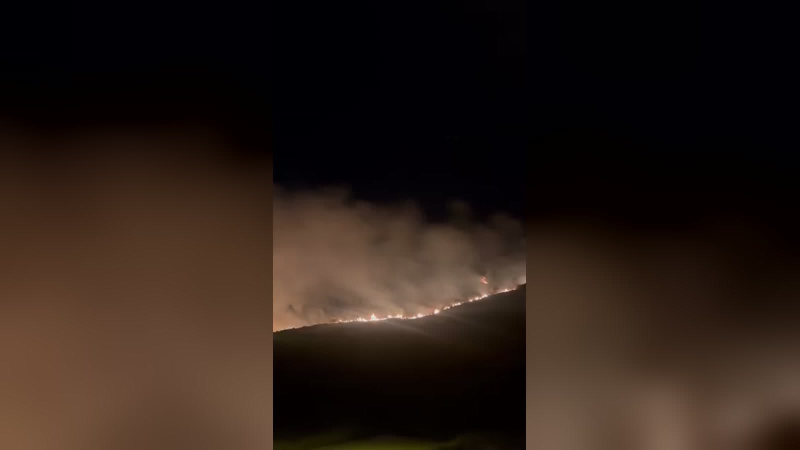 De noche, una línea de fuego avanza por la ladera de un monte y levanta una gran nube de humo iluminada de naranja.