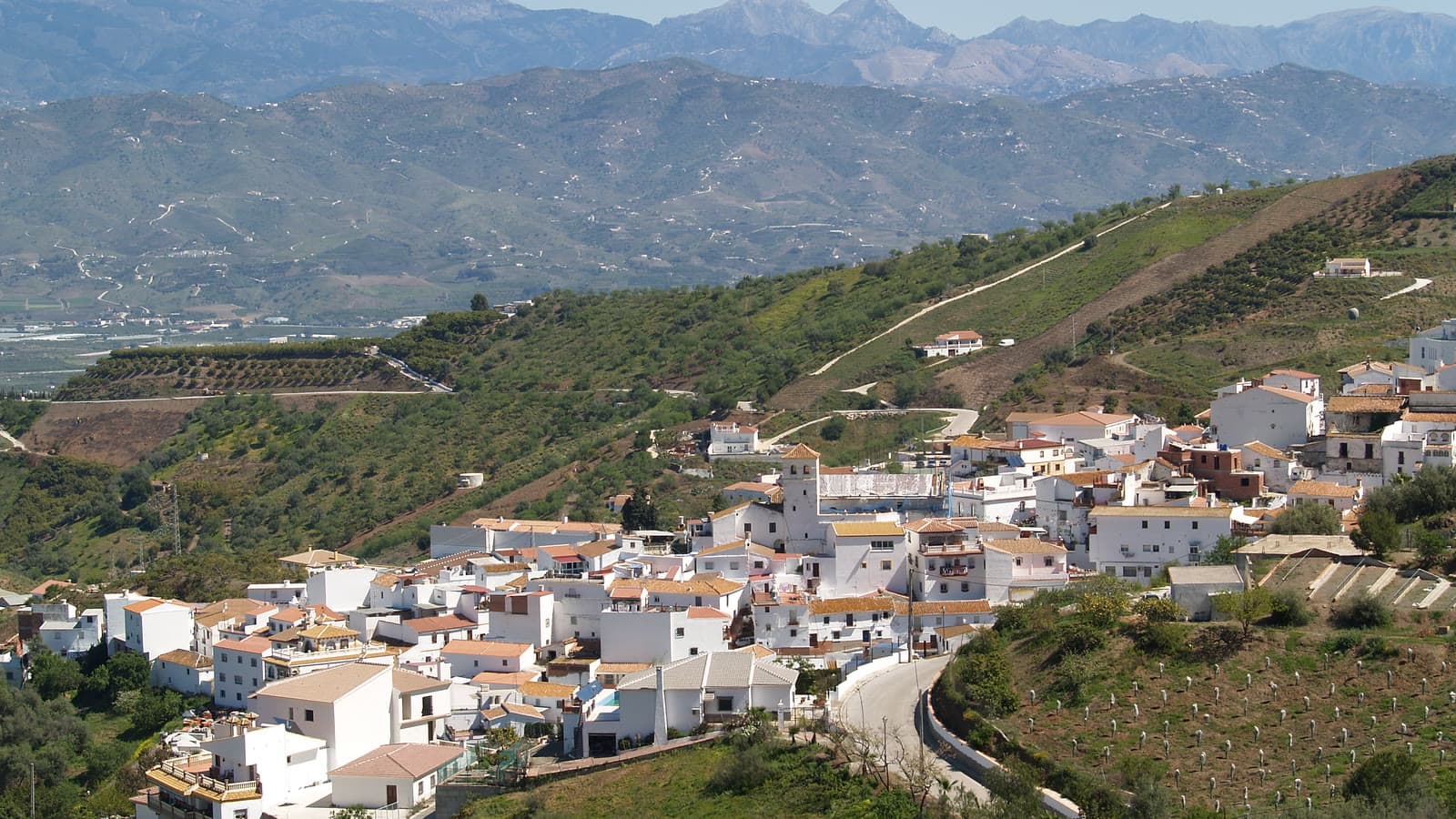 Vista de Iznate: casas blancas en una ladera con la torre de la iglesia en el centro, campos de cultivo alrededor y montañas al fondo.