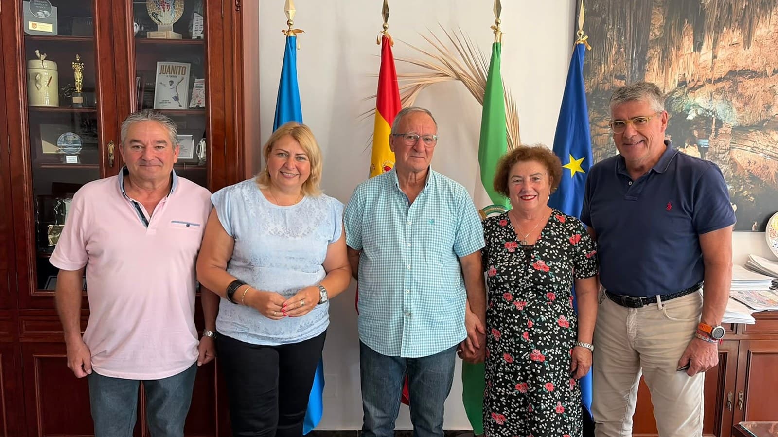 Cinco personas posan sonrientes ante las banderas en un despacho del Ayuntamiento de Nerja; entre ellas, María del Carmen Centurión, con un vestido negro de flores rojas.