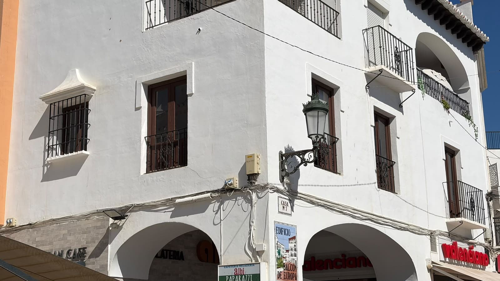 Fachada blanca del edificio Balcón de Europa de Nerja, con balcones de forja, una farola y soportales con comercios en la planta baja.