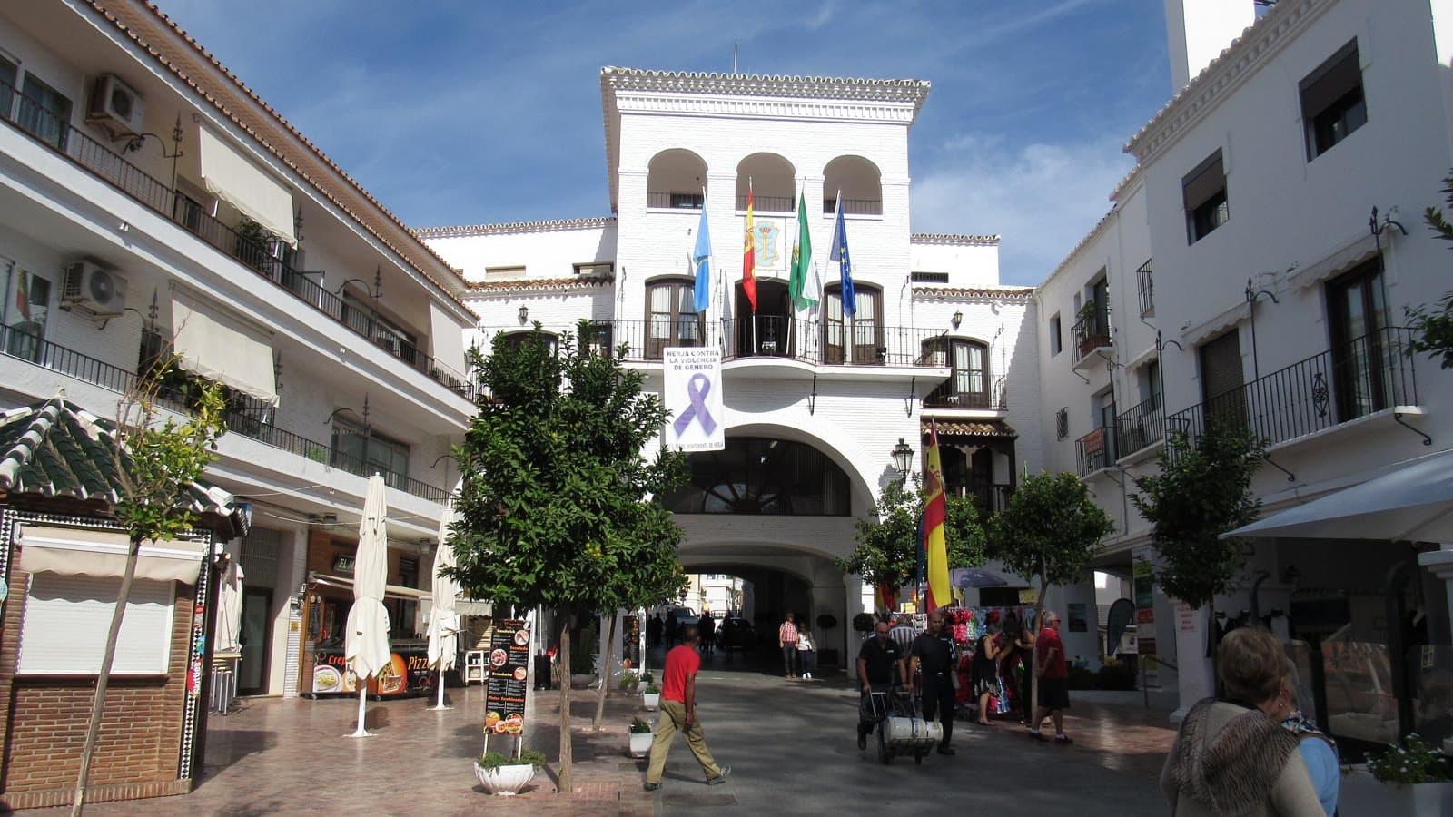 Fachada blanca del Ayuntamiento de Nerja con arcos, balcón de forja y banderas, vista desde una calle peatonal con naranjos y terrazas.