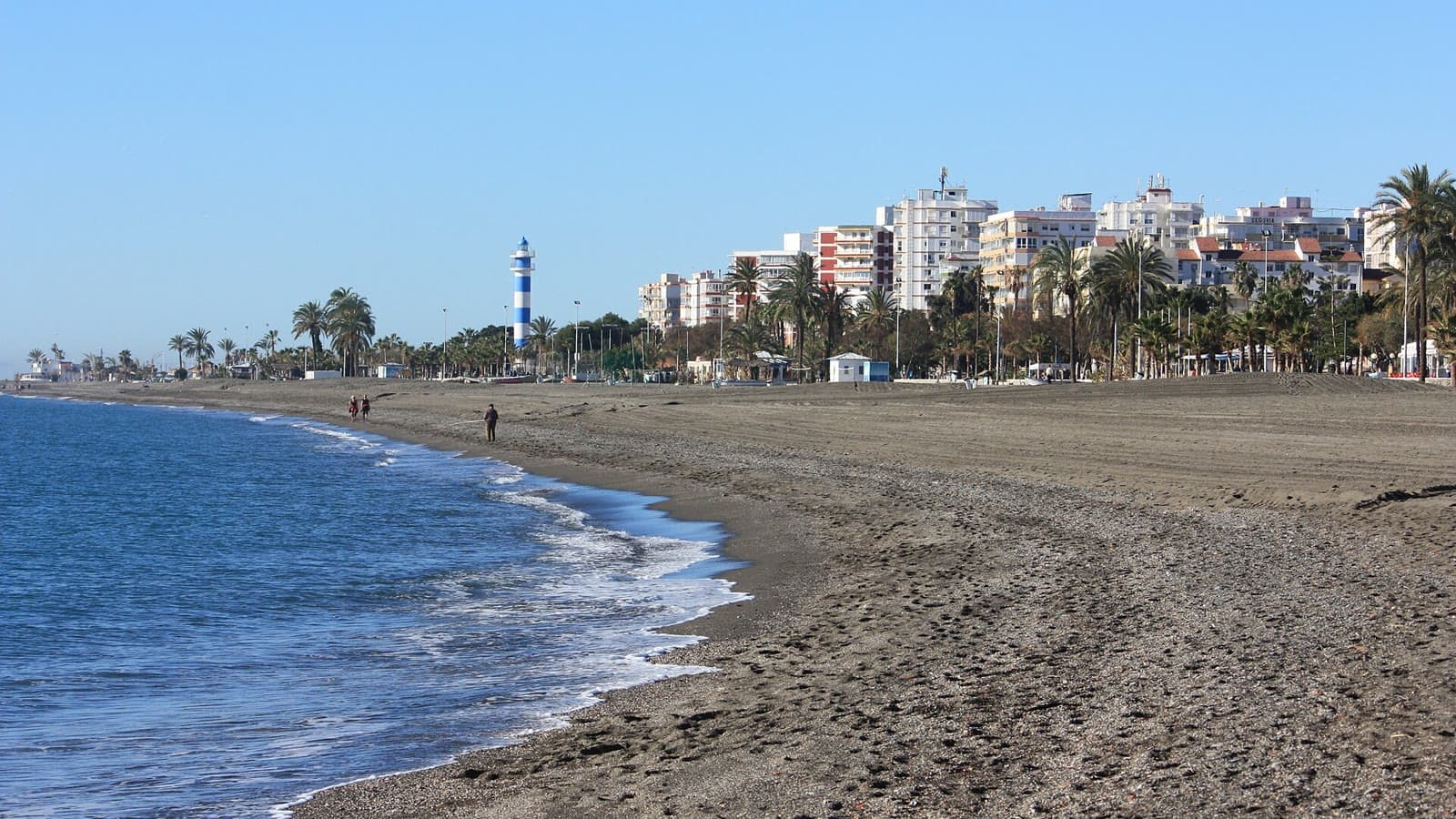 Playa de arena oscura de Torre del Mar con el faro de rayas azules y blancas, palmeras y edificios del paseo marítimo al fondo.