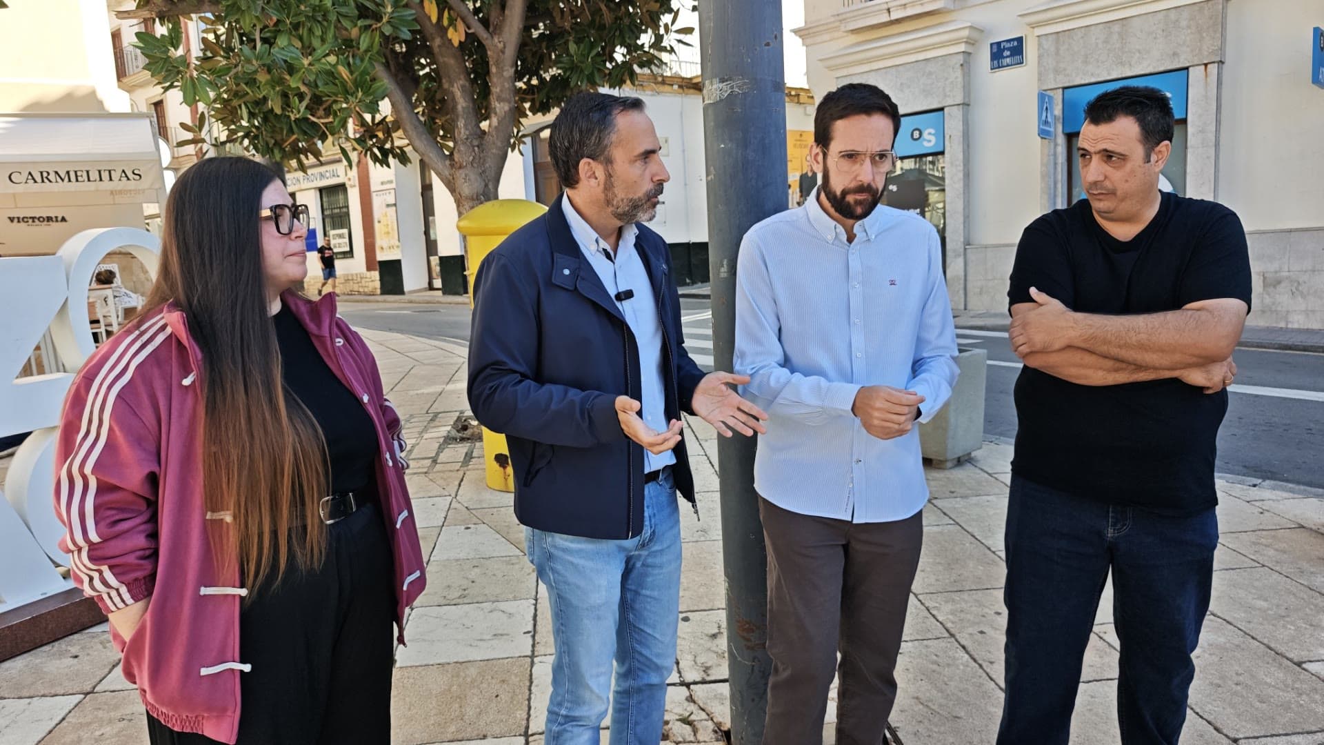 Cuatro personas conversan de pie en la plaza de las Carmelitas de Vélez-Málaga, junto a una farola y un árbol.