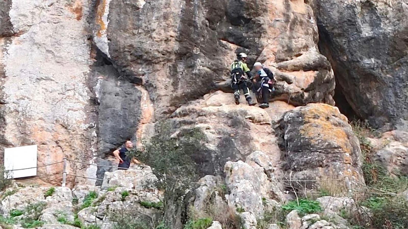 Dos bomberos con casco y arnés junto a una persona en una pared de roca de la vía ferrata de Comares, con otro hombre más abajo en el sendero.