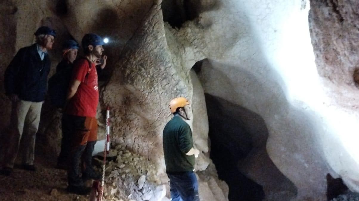 Cuatro personas con casco observan una gran formación de roca blanquecina iluminada en el interior de la Cueva de la Victoria
