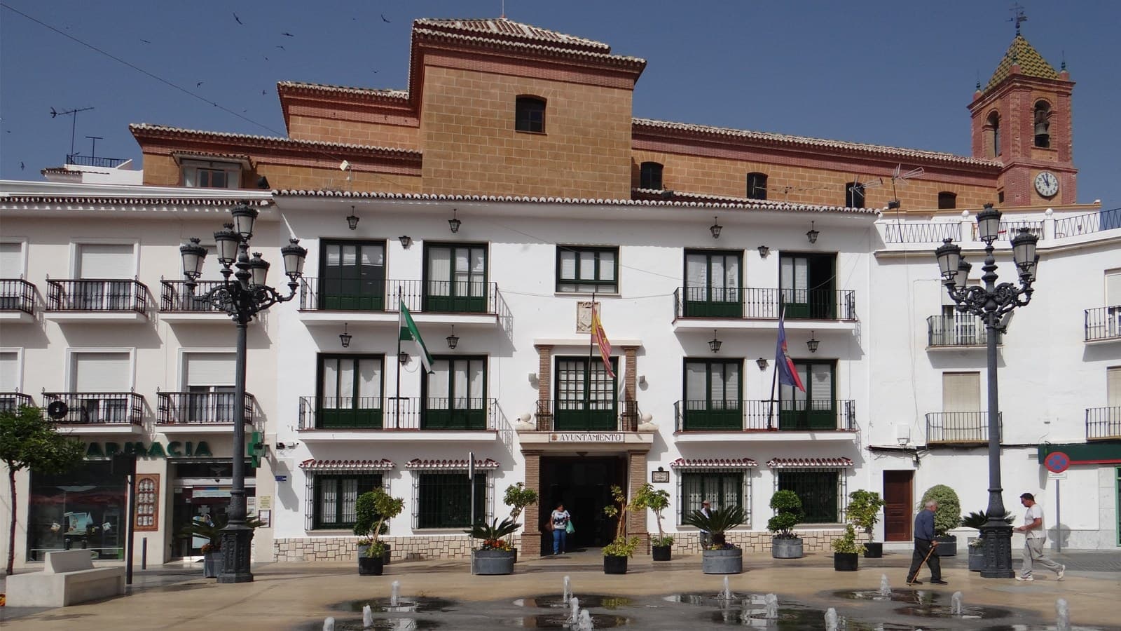 Fachada blanca del Ayuntamiento de Torrox, con balcones de rejería y ventanas verdes, banderas sobre la puerta y un cartel que dice «Ayuntamiento»; detrás asoma un edificio de ladrillo y la torre del campanario, y delante una plaza con farolas y fuentes a ras de suelo.
