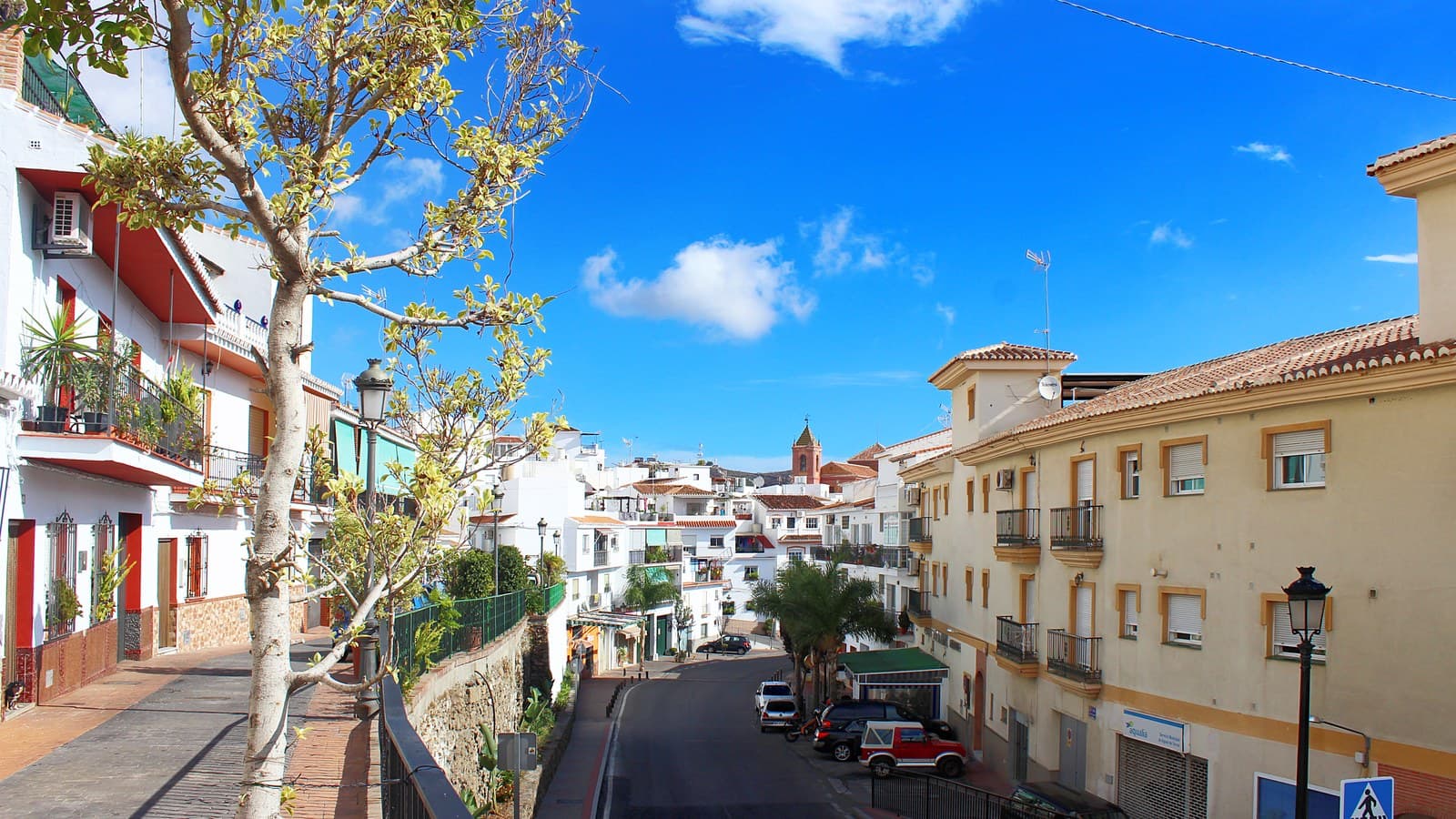 Calle en cuesta de Torrox pueblo bajo un cielo azul con nubes, entre casas blancas con balcones a la izquierda y un bloque de color crema a la derecha; al fondo, entre los tejados, asoma el campanario de la iglesia.