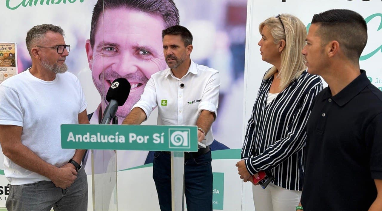 José Pino, con camisa blanca de Andalucía Por Sí, habla tras un atril del partido ante un micrófono, acompañado de tres miembros de su equipo, delante de un cartel con su foto.