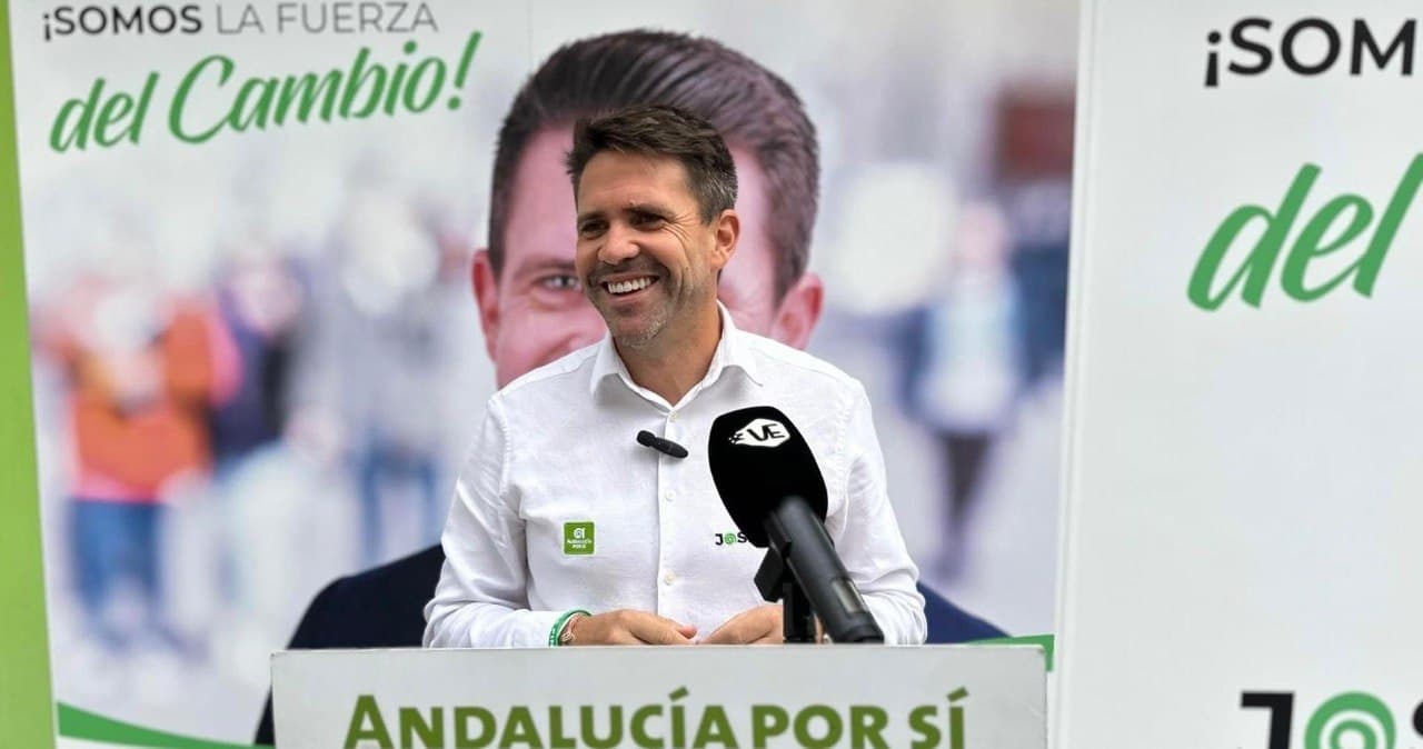 José Pino, sonriente, con camisa blanca y la insignia de Andalucía Por Sí, habla tras un atril del partido ante un micrófono, con un cartel de «¡Somos la fuerza del cambio!» al fondo.