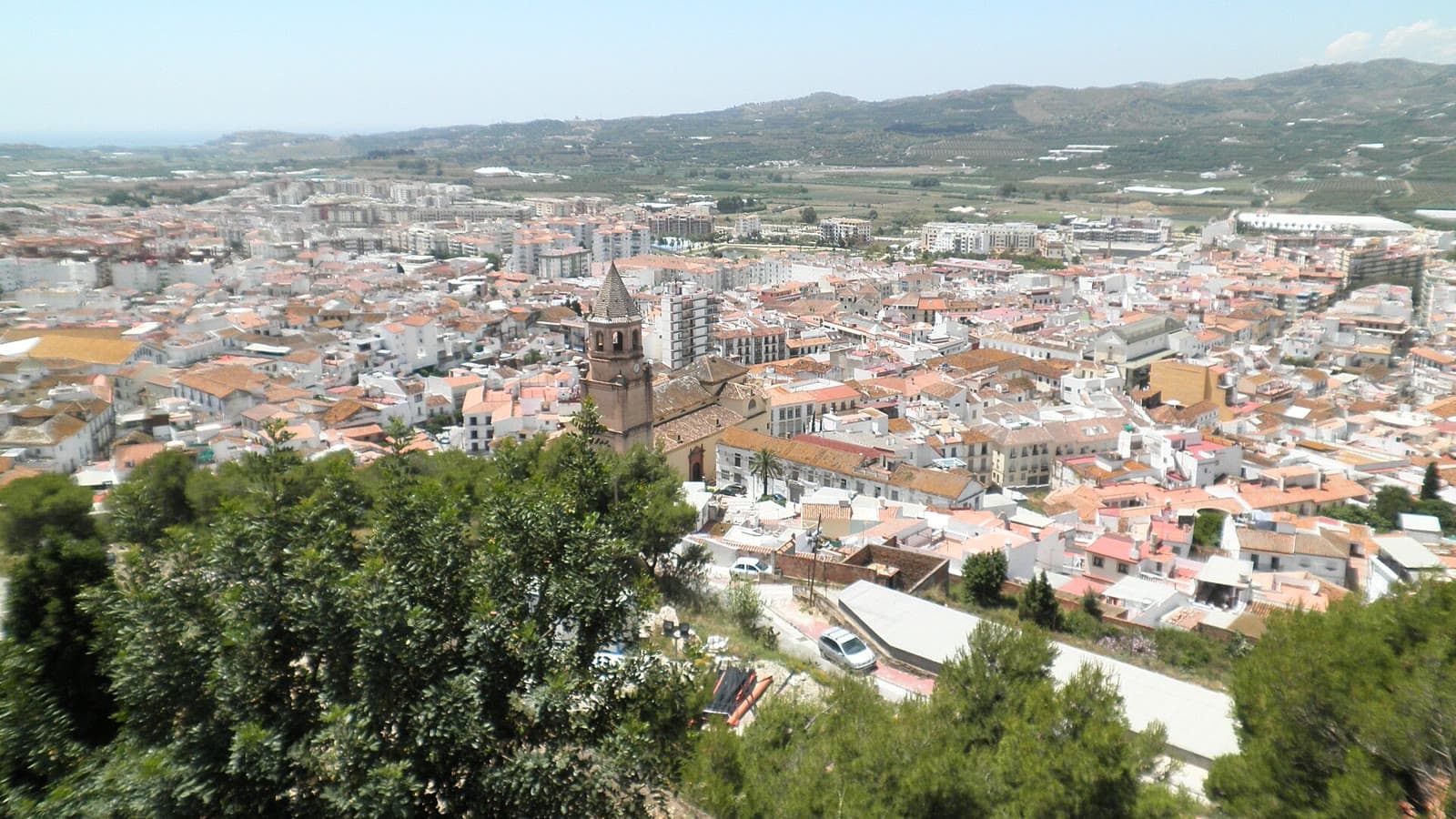Vista desde lo alto de Vélez-Málaga con los tejados del casco urbano, una torre de iglesia en primer plano y la vega y los montes al fondo
