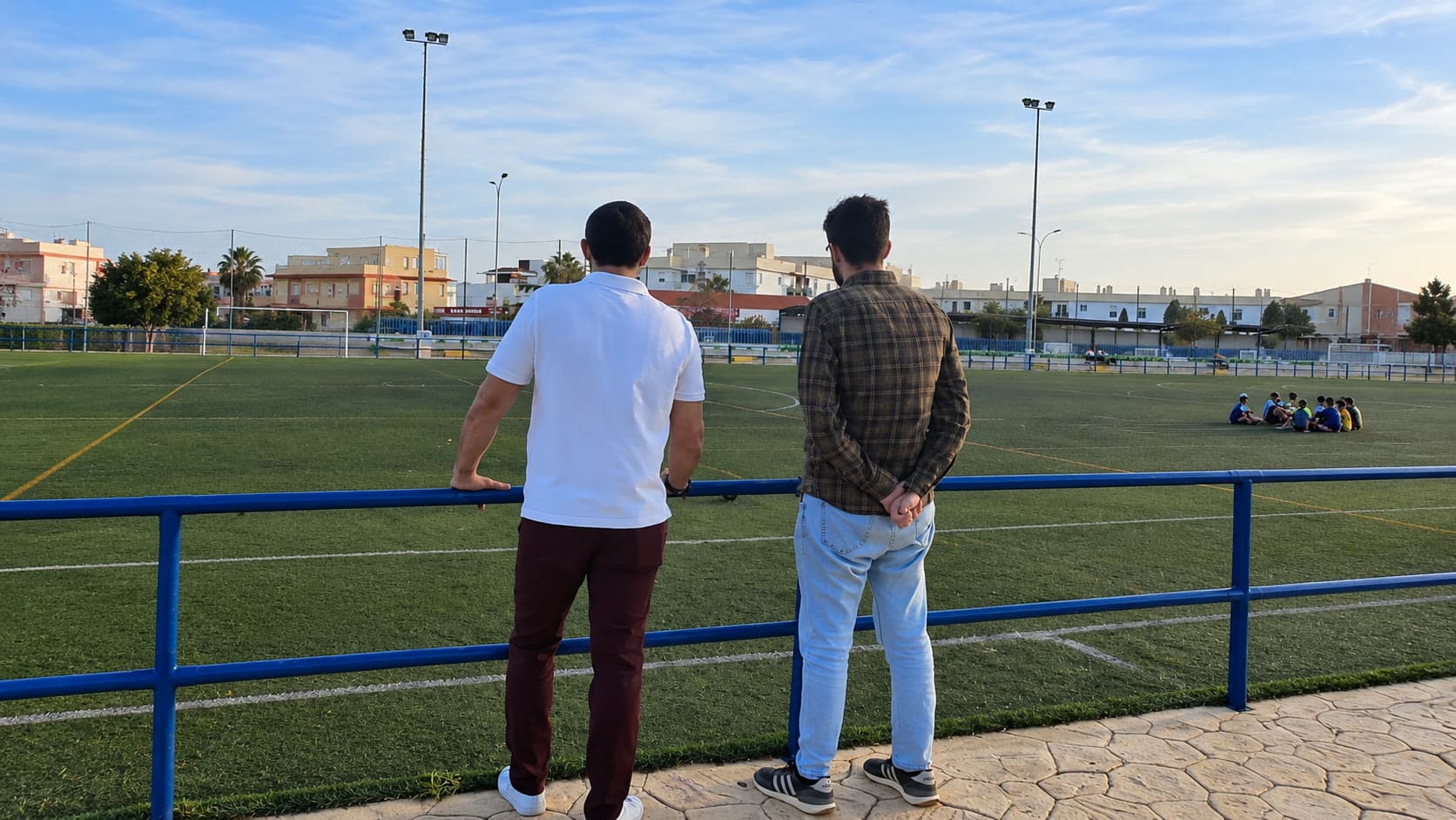 Dos hombres, de espaldas, apoyados en la barandilla azul del campo de fútbol de césped artificial del polideportivo Fernando Ruiz Hierro; al fondo, un grupo de niños sentados en el campo y edificios de Vélez-Málaga.