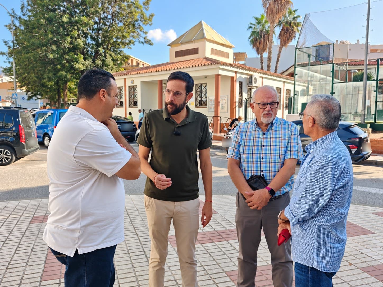 Cuatro hombres conversan en una acera de Vélez-Málaga, delante de un edificio municipal de una planta con tejado de teja y una pista deportiva vallada; detrás hay coches aparcados.